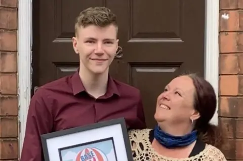 Danny Donnelly Danny Donnelly pictured standing next to his mum whilst holding a police award he received. Danny is wearing a burgundy shirt, his mum is wearing a beige crochet jumper. 