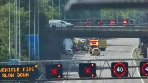 National Highways A highways camera view of work gire and police near a lorry on a hard shoulder. Gantries are showing warning signs.