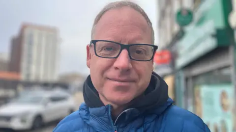 Paul Berry smiling at the camera is wearing black framed glasses and a blue puffer jacket.