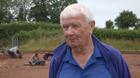 John has white hair and a blue T-shirt and jumper. He stands in front of people digging in a field.