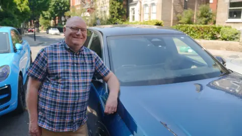 Gwyndaf Hughes/BBC News A smiling man in a checked blue and red shirt, and brown trousers leans on his dark blue car parked in the middle of the street. To his right is a lighter blue car and in the background a residential street with other cars driving both ways and front garden hedgerows along the pavement