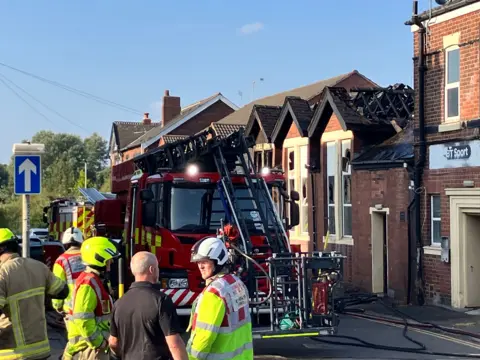 Firefighters wearing light brown uniforms and high-vis jackets and helmets stand in a street with a fire engine beside a single-story building with damaged and blackened roof gables. The damaged building is behind a grander, red-brick, two-story building.