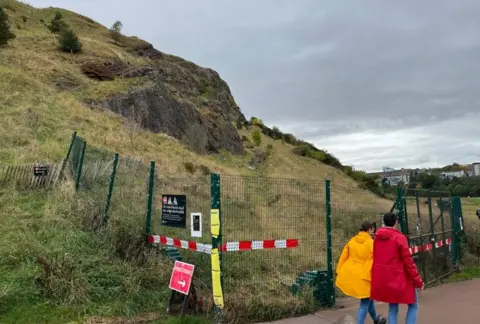 Two people in red and yellow jackets walk past a green fence that is keeping people out of the Radical Road Path in Holyrood Park. 