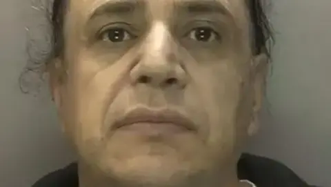 A mugshot of a man with dark brown eyes and long dark hair that is pulled back off his face. He is staring into the camera and the background is grey.