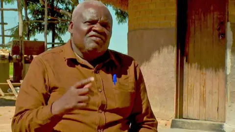 Godfrey Gurira sitting on a chair in front of a small round and thatch building. He is wearing a brown long sleeved shirt and gestures with his finger as he talks