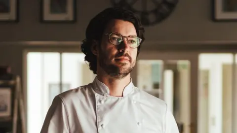 Kent Cookery School a man with glasses and curly black hair stands in a kitchen wearing his chef whites