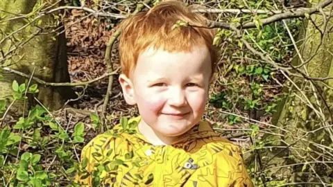 Family handout George with ginger hair, wearing a yellow t-shirt standing in woodland smiling. 