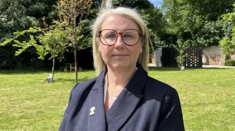 George Carden/BBC A woman with large glasses and long, blonde hair looking at the camera. She is wearing a black blazer and is standing in a large garden.