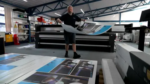 Andy Meeson/BBC A man wearing shorts and a dark blue T-shirt carries a large sheet of white, printed card towards a pile of similar material stacked on the floor, in the foreground of the image. The man has a greying beard and grey hair. He is striding purposefully from a large, industrial printer in the background and towards the pile of printed card on the floor. The image is taken inside a commercial facility, with dark carpets, strip lighting and white walls.