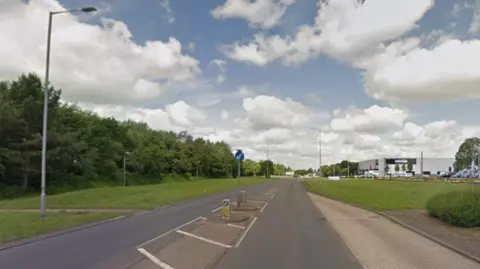 Google A stretch of single carriageway road with a lay-by on the right and a streetlight on the left. There are grass verges on either side of the road and a large grey building in the distance. 