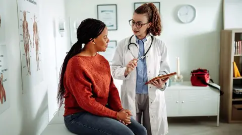 Woman with long black hair sits on a doctor's examination table listening to a female doctor wearing a white coat and stethescope. 