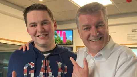 Jack Soley smiling at the camera in a blue GAP hoodie. He stands next to BBC Radio Wiltshire presenter Graham Rogers, also smiling and wearing a white shirt. 