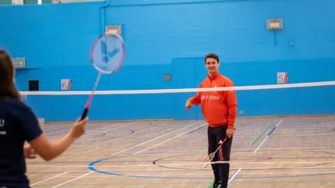 University of Bristol Dan in a red tracksuit top and black bottoms, playing badminton with a coach on an indoor court