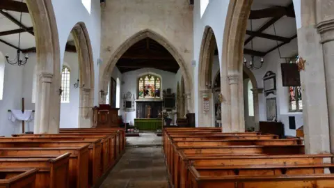 The interior of a medieval church. There are pews on either side and arches. In the distance of an alter with a stained glass window above.