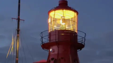 The top of a lighthouse is lit up at dusk. It's glowing with an orange light and a ship's mast can be seen to the left of it. The sky is cloudy and a dark blue-grey colour.