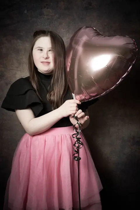 Debbie Todd Ava, an older girl with long brown hair wearing a black top and pink skirt holds a large pink heart-shaped balloon.