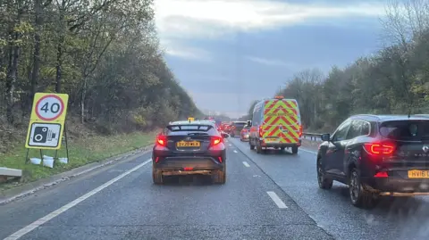 Shaun Whitmore/BBC Vehicles queuing on a section of dual carriageway on the A11. The picture shows the backs of a number of vehicles and a yellow sign with a 40mph speed limit.