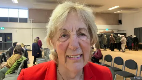 Head and shoulders shot of Fay Gorwood standing inside a community centre. She is wearing a red jacket and smiling at the camera. There are a number of people in the background and rows of chairs.