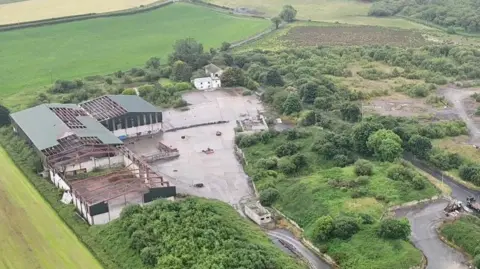 The site of an illegal dump sits surrounded by green fields and countryside. a number of dilapidated buildings are on the left, while minor roads run across the width of the site