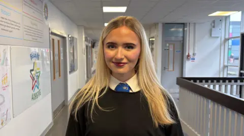 Secondary school pupil Eve. She has long blonde hair and is wearing a white shirt, blue tie and black jumper. She is standing in the school corridor, where there are infographics on the wall and doors to classrooms in the background.