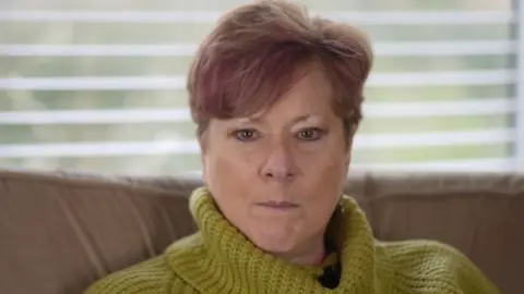 Lynne O'Neill has short hair and looks directly into the camera. She is wearing a green roll-neck sweater and sitting on a brown sofa with a white Venetian blind behind her.