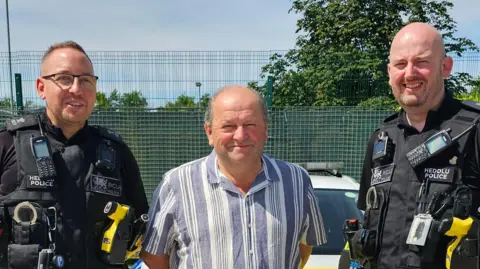 North Wales Police Neville, an older man with a blue and white stripped shirt with two polices officers in uniform on either side of him. 