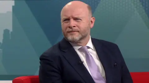 Liam Byrne MP sits in the BBC Politics Midlands studio, wearing a black suit and lilac tie