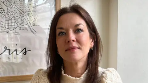 Delphine Pesrin-Auder - A woman with dark hair and blue eyes looks at camera from inside her cafe, with a poster that says "Paris, France" in the background.