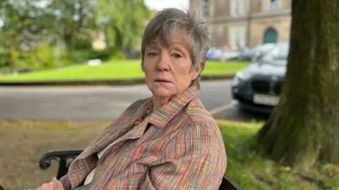 Woman sat on park bench with short grey hair and multi coloured jacket