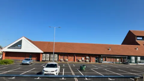 A large red brick building, with a red tiled roof. There are NHS signs to the left and right, and a large car park in the foreground 