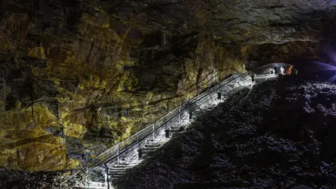 An underground tunnel with steps leading down. It is dark but the steps have white lights on them. The walls are solid rock. 