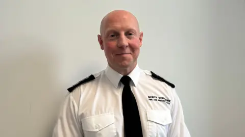 BBC/Emily Johnson A man smiles at the camera wearing a North Yorkshire Fire and Rescue Service branded white shirt and black tie. He is clean shaven and has a bald head.