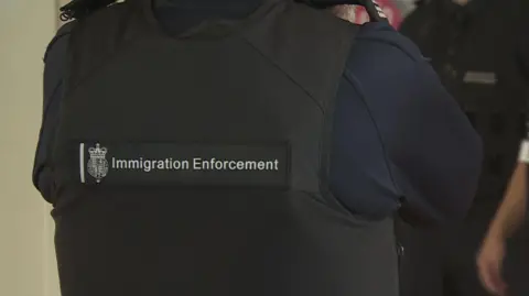 BBC An Immigration Enforcement serviceman  lasting  with their backmost  to the camera. They are wearing a achromatic  vest with a 'Immigration Enforcement' spot  connected  the back.