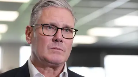 Sir Keir Starmer has a serious expression and is looking at the camera. He has short grey hair and is wearing black-framed glasses, a black suit and white shirt.