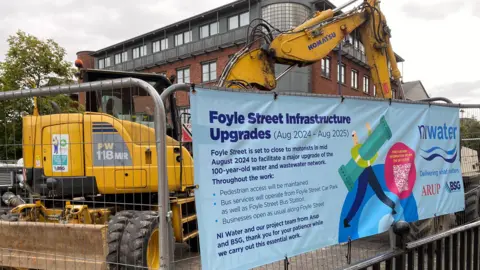 BBC Barriers closing off Foyle Street , a digger is behind, and a sign is tied to the barriers notifying the public of the sewerage works