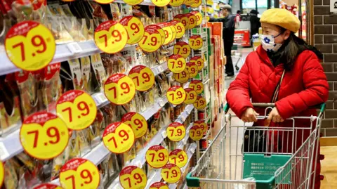 A customer wearing a mask and winter jacket browses through products at a supermarket in Lianyungang, in eastern China's Jiangsu province on January 9, 2025. She is standing in an aisle with a trolley in front of her, flanbked by shelves with yellow-colour discount promos on them. 