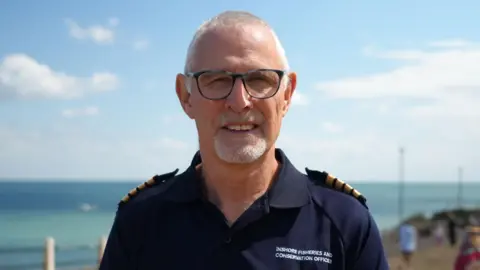 Shaun Whitmore/BBC Julian Gregory standing outdoors at the coast. The sea is in the background behind him. He is looking directly at the camera and has a pair of circular glasses frames on his eyes. 