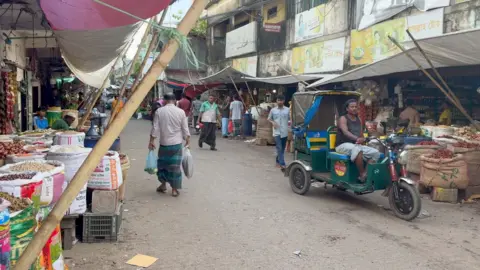 UNHCR A busy outdoor market scene with stalls selling various goods, including large sacks of dried chilies, onions, and other produce. People walk along the street carrying shopping bags, and a green auto-rickshaw with a driver is parked on the right side. Makeshift tarps provide shade over the stalls, and signs with text in Bengali are visible on the buildings.