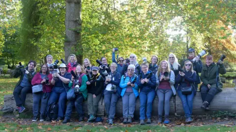 A group of women smiling for the camera on a sunny day outside. They are holding cameras. They are sitting on the trunk of a big tree.