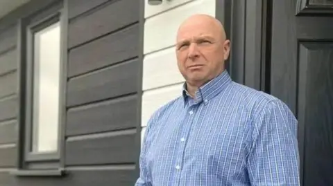 Mike Chesworth, wearing a blue checked shirt, stands with his hands in his pockets in front of the door of his dark grey lodge