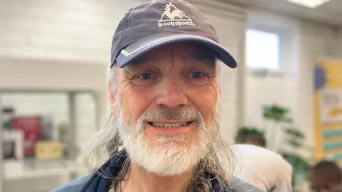 Stephen Smith, who has a white beard and long hair, smiles for a photo wearing a blue sports cap at one of the digital inclusion sessions. 