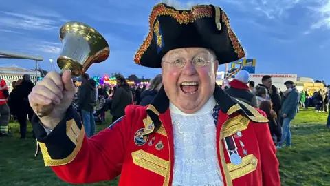A tow crier, in full uniform of red jacket an black hat, raises a bell
