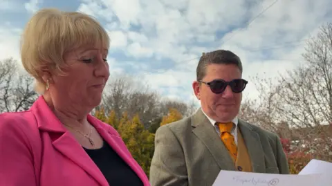 A woman with cropped blonde hair in a pink jacket and a man in a jacket and waistcoat with dark glasses reading letters in a garden.