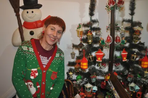Lisa Watts, a smiling woman in a green Christmas cardigan leans beside a small decorative Christmas tree adorned with colourful lantern ornaments, with a snowman figure behind her and a mirror reflecting the festive display.