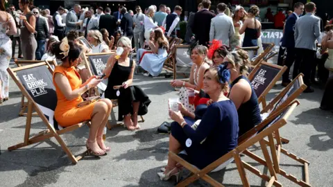 PA Media Six women in formal dresses, three with fascinators, sit on a collection of black deckchairs near a crowd of racegoers