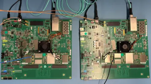 Ben Schofield/BBC Two large, square printed circuit boards, each filled with electronic components and with wires connected to and from them. The shot is taken from above, looking down on the two boards, side-by-side, laying on a flat, light blue surface. The boards themselves are deep green in colour, while the metal components are catching the light from above.