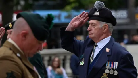 Two men in military uniform. A man in a navy blazer with medals and a hat salutes.