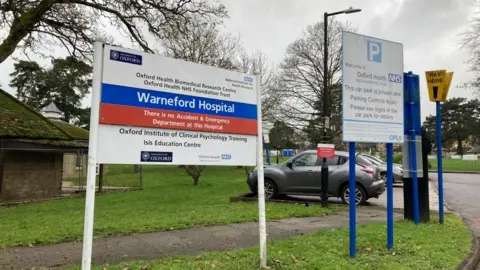 A sign outside a hospital saying Warneford Hospital. There's a carpark in the background and some autumnal trees.
