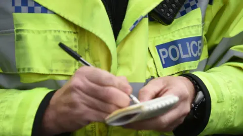 A police officer wearing a yellow high visibility jacket is writing in a notepad. There is a blue logo on the jacket identifying the person as a police officer. They are also wearing a watch, visible under the sleeve of the hand holding the notepad.