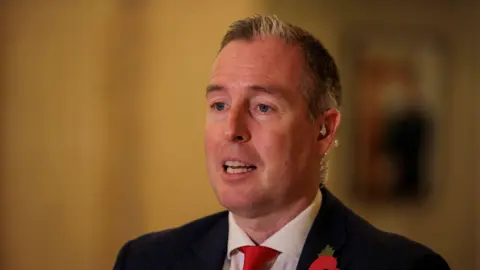 Paul Givan speaking. He is wearing a black suit jacket, white shirt and red tie.